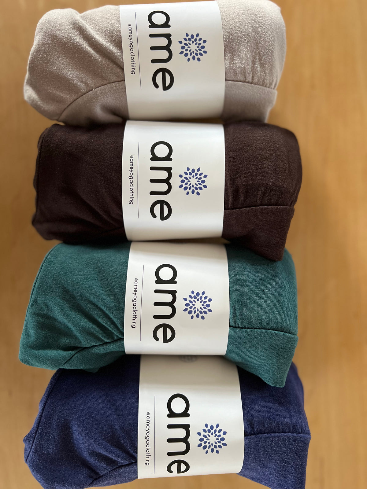 Four pairs of shorts in gray, brown, green, and blue with 'ame' branding on a wooden surface.