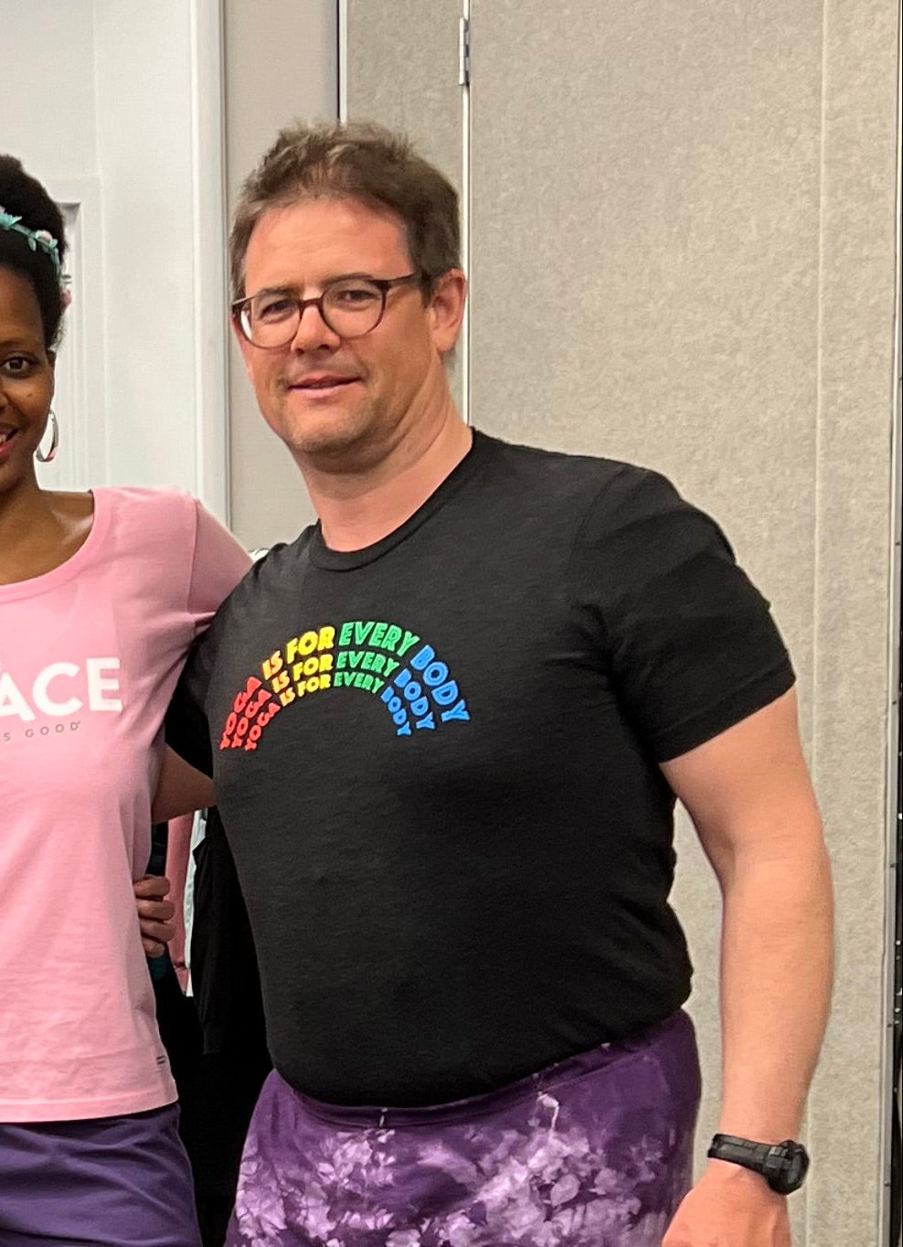 A tall gentleman wearing a black tshirt with a rainbow graphic that says "Yoga Is For Every Body" and purple tie dye yoga shorts. 