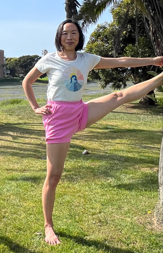 One woman posing outdoors in a park on a sunny day, performing a yoga stretch on the grass with palm trees and blue sky in the background.